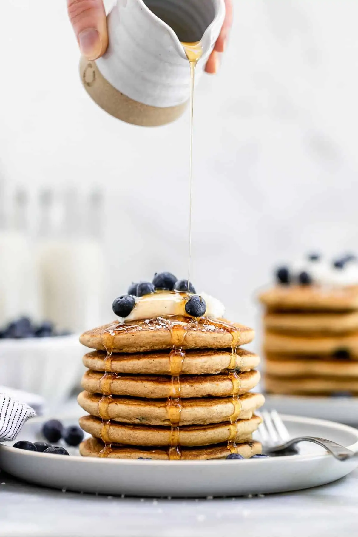 Two plates with pancakes stacked on each other with blueberries and maple syrup pouring on top.