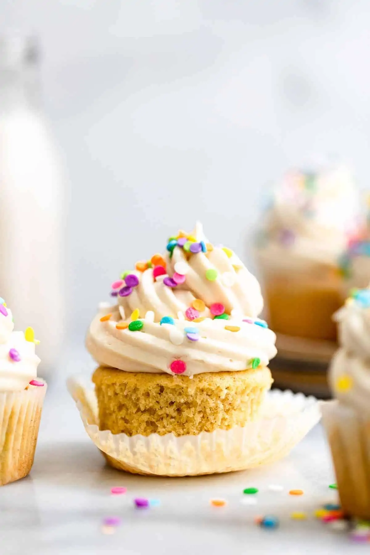Gluten free vanilla cupcakes with rainbow sprinkles on top with milk in the back.