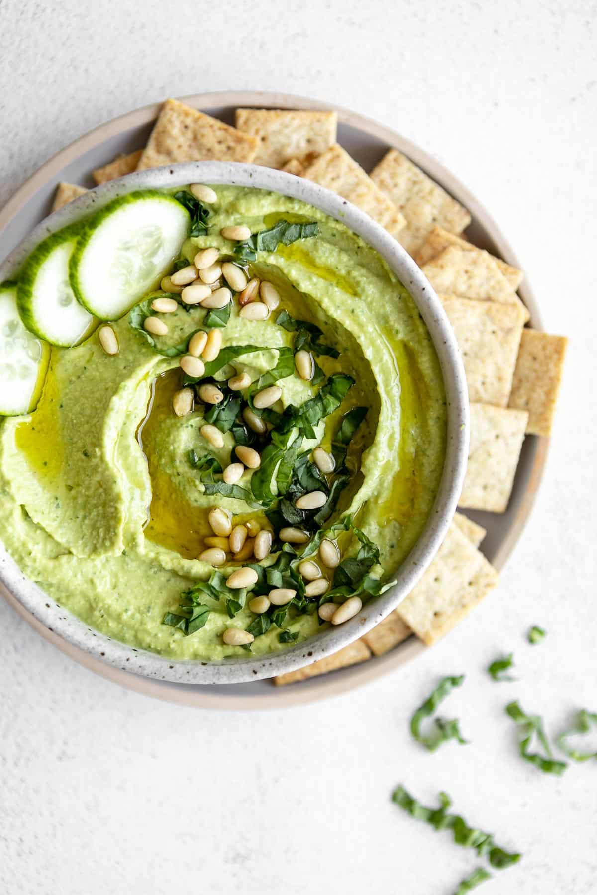 pesto hummus in a bowl with crackers and cucumber on the side